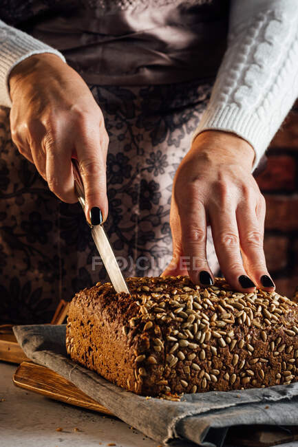 Анонимная пекарь с ножом режет буханку свежего хлеба с семечками подсолнуха на столе в пекарне — стоковое фото