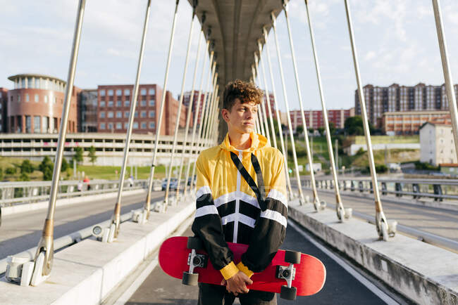 Красивый белый мальчик со скейтбордом, стоящий посреди моста. — стоковое фото