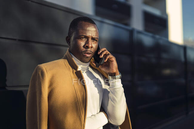 Concentrado jovem afro-americano masculino em gola alta elegante e casaco falando no telefone celular e olhando para longe pensativo enquanto estava na rua da cidade — Fotografia de Stock