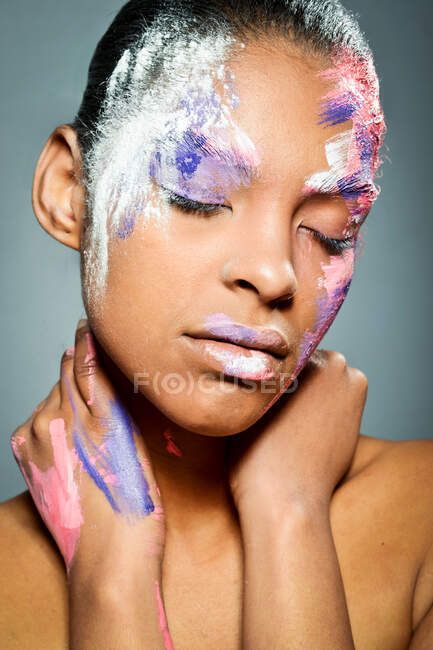 Modèle féminin ethnique créatif avec visage enduit de peinture rose et blanche touchant le cou avec les yeux fermés sur fond gris en studio — Photo de stock