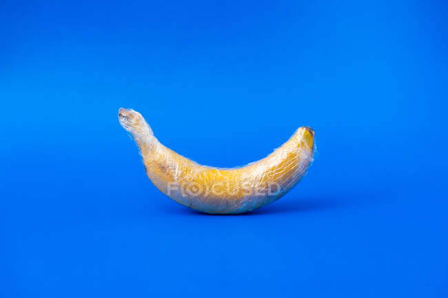 Смачний стиглий банан, покритий прозорою пластиковою обгорткою, що представляє концепцію промислового сільського господарства на яскравому фоні — стокове фото