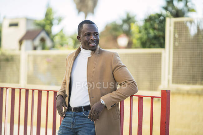 Happy young ethnic male in trendy outfit leaning on a fence of street on sunny day — Stock Photo