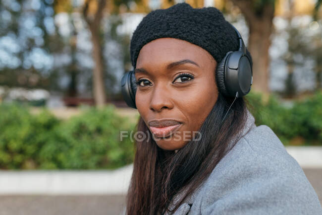 Acercamiento mujer afroamericana en ropa de abrigo sentada en la calle y escuchando música en auriculares - foto de stock
