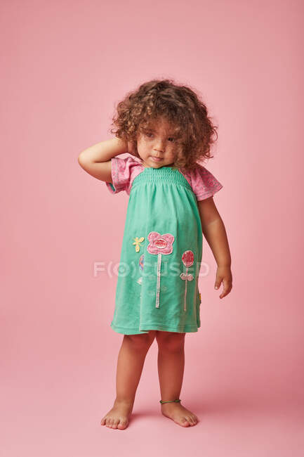 Criança adorável em vestido com cabelo encaracolado olhando para longe de pé no chão de fundo rosa — Fotografia de Stock