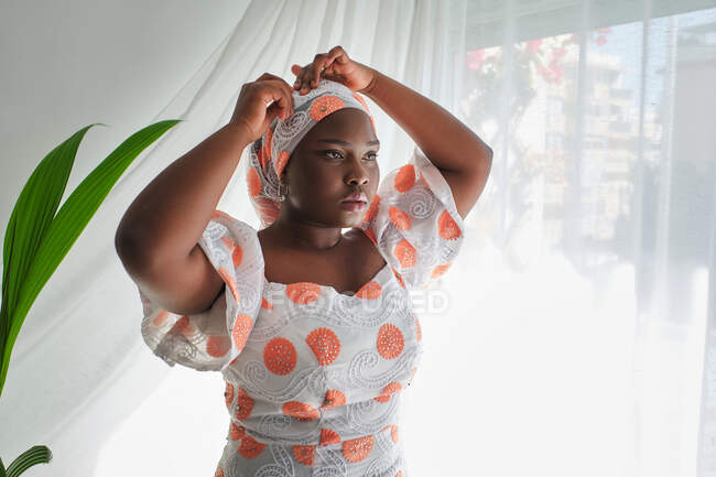 Concentrated young African lady in stylish summer dress adjusting traditional turban while standing near window in light room — Stock Photo
