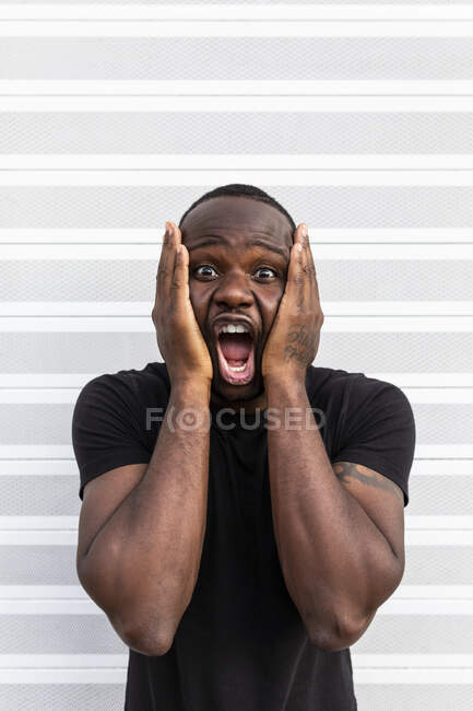 Giovane stupito maschio afroamericano in t shirt nera gridando mentre si tocca il viso e guardando la fotocamera su sfondo chiaro — Foto stock
