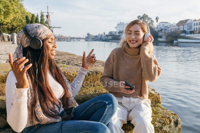 Positive multirassische Freundinnen, die auf einem Damm sitzen und mit geschlossenen Augen Lieder mit Kopfhörern genießen, während sie zusammen Spaß haben — Stockfoto