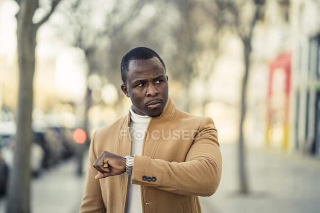 Концентрированный молодой афроамериканец в модной одежде проверяет время на наручных часах во время прогулки по городской улице в солнечный день — стоковое фото