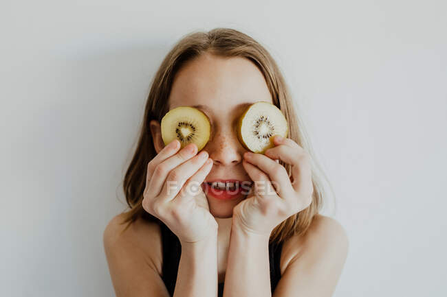 Chica alegre en ropa casual sonriendo mientras cubre los ojos con rodajas de kiwi sobre fondo blanco - foto de stock