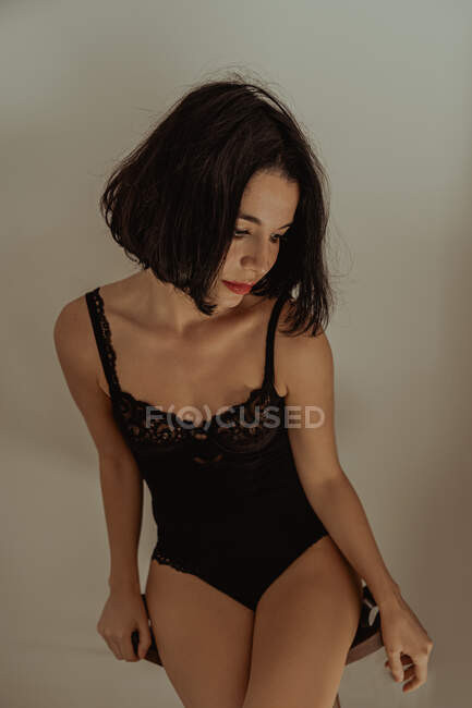 Serene female in black lace bodysuit sitting on stool in room against white wall and looking away — Stock Photo