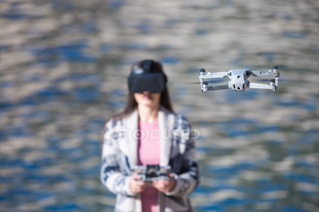 Aufgeregte Frau mit moderner VR-Brille bedient Drohne mit Fernbedienung und erlebt Virtual Reality, während sie vor verschwommenem Meer steht — Stockfoto