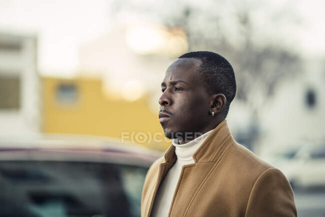 Side view of portrait of confident young ethnic male in trendy outfit walking on city street and looking away on sunny day — Stock Photo