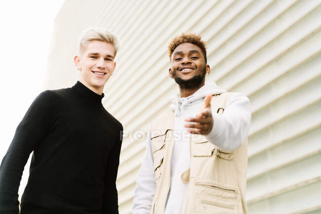 From below happy young multiracial male friends in trendy outfits standing on city street near modern building and looking at camera — Stock Photo