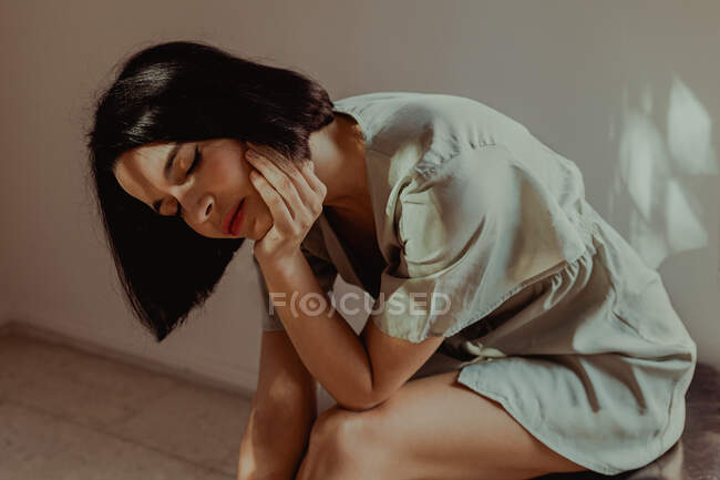 Side view of tender female leaning on hands while sitting with closed eyes on stool in room lit by sunlight — Stock Photo