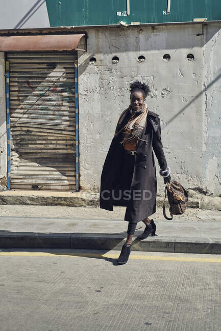 Giovane donna afroamericana felice in abiti alla moda con auricolari che camminano su strada asfaltata mentre guarda la fotocamera in città — Foto stock