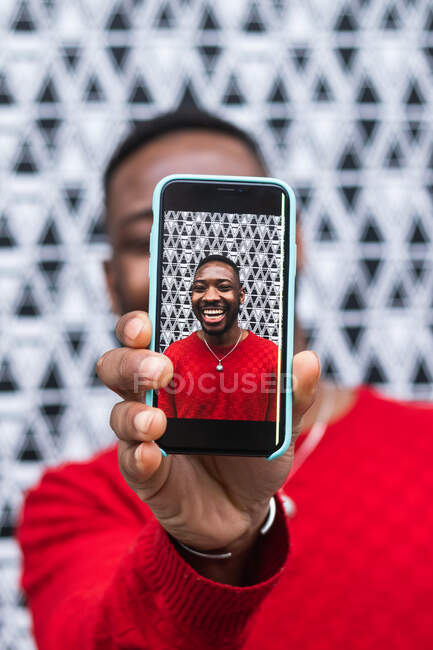 Hombre étnico en ropa brillante que cubre la cara con el teléfono celular mientras demuestra la foto en la pantalla durante el día - foto de stock