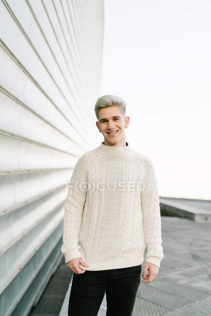 Vista laterale di giovane ragazzo alla moda in maglia maglione alla moda in piedi sulla strada della città vicino a edifici contemporanei nella giornata di sole — Foto stock