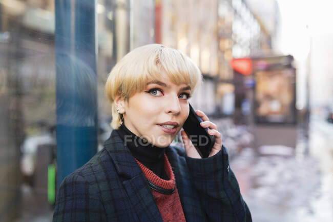 Positive young female with short hair in warm coat having conversation via mobile phone while standing on snowy city street and looking away in Madrid, Spain — Stock Photo
