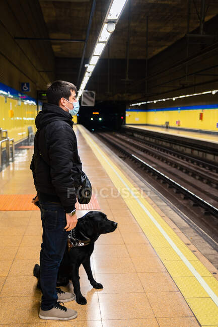 Вид сбоку под низким углом все тело анонимного слепого мужчины в маске стоящего с собакой-поводырем на поводке на платформе — стоковое фото