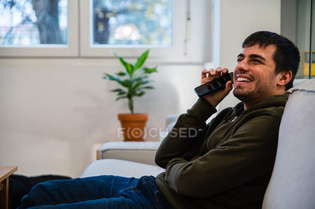Вид збоку молодого усміхненого чоловіка, який страждає від сліпоти, сидить на дивані і слухає аудіоповідомлення на мобільному телефоні — стокове фото