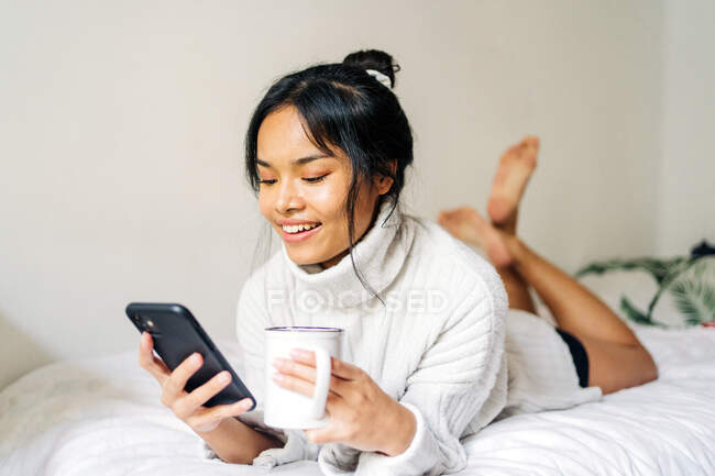Capelli lunghi bruna donna asiatica sdraiata sul letto a casa e utilizzando un cellulare — Foto stock