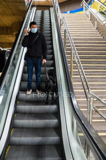 Corpo completo di maschio in maschera con disabilità visiva in piedi su scala mobile con cane guida e utilizzando il telefono cellulare — Foto stock