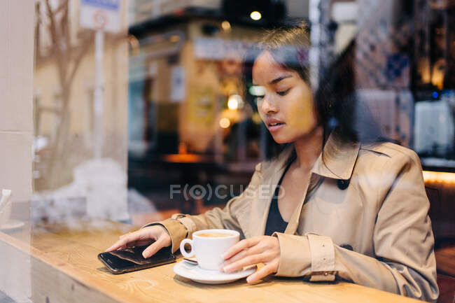Brune aux cheveux longs femme asiatique prendre un café sur un café tandis que cherche un téléphone portable — Photo de stock