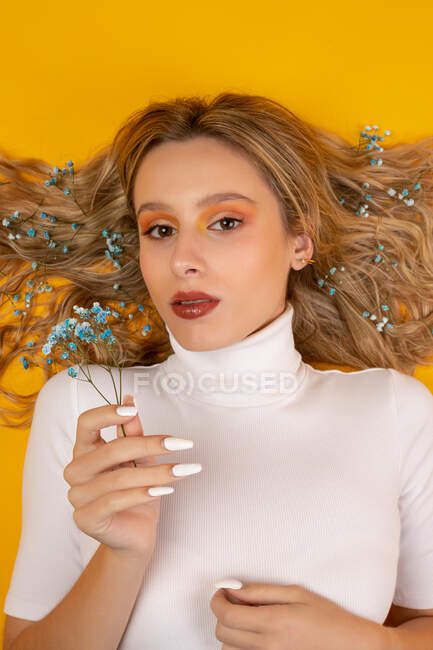Attractive young female lying down on floor with blue tender gypsophila flowers on the hair on yellow background in studio looking at camera — Stock Photo