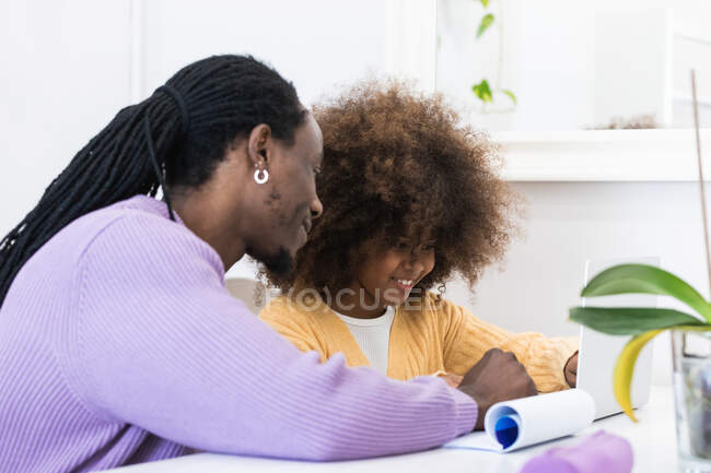 Sorrindo pai afro-americano sentado à mesa com menina surfar netbook enquanto prepara tarefa para a escola — Fotografia de Stock