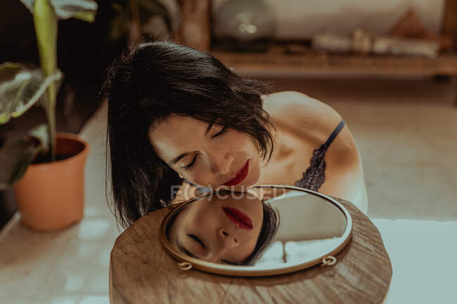 Mujer pacífica sentada con los ojos cerrados en el suelo en la habitación y reflejándose en el espejo de forma redonda - foto de stock