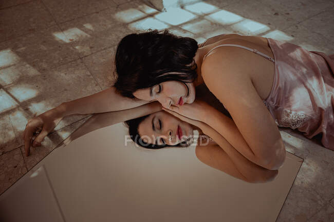 Alto angolo di tenera donna in camicia da notte sdraiato con gli occhi chiusi sul pavimento e riflettente nello specchio — Foto stock