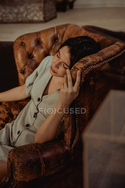 From above of peaceful female sitting in shabby leather chair and touching head while relaxing at home — Stock Photo
