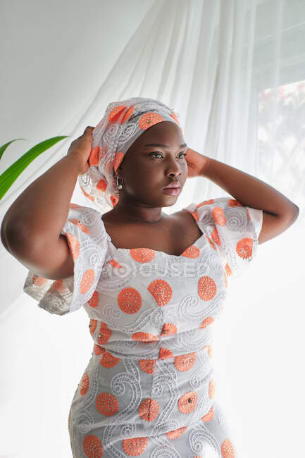 Jovem senhora africana concentrada em vestido de verão elegante ajustando turbante tradicional enquanto está perto da janela na sala de luz — Fotografia de Stock