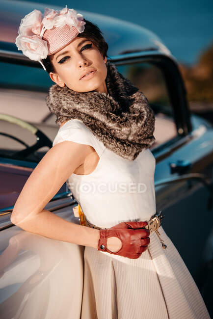 Corpo inteiro de mulher elegante vestindo luvas de couro vestido longo e chapéu de pé perto do carro vintage contra o mar — Fotografia de Stock