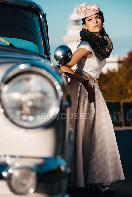 Corpo inteiro de mulher elegante vestindo luvas de couro vestido longo e chapéu de pé perto do carro vintage contra o mar — Fotografia de Stock