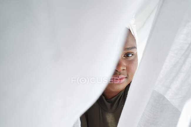 Positive young African female in traditional headscarf hiding behind white curtains and looking at camera in sunlight — Stock Photo
