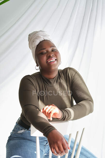 Happy alluring young black lady in casual clothes and turban sitting on chair and looking at camera — Stock Photo