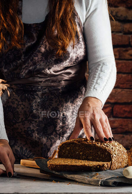 Crop boulanger femelle anonyme avec pain frais coupé au couteau avec des graines de tournesol sur la table dans la boulangerie — Photo de stock