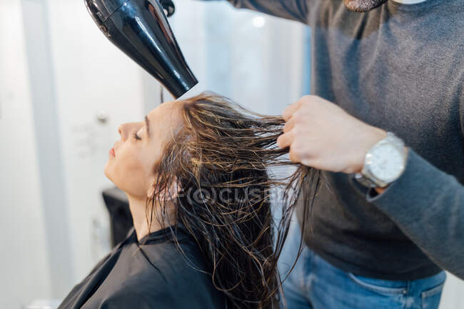 Растениеводство молодой этнический мужчина парикмахер сушки волос клиентки с закрытыми глазами в современной студии красоты — стоковое фото