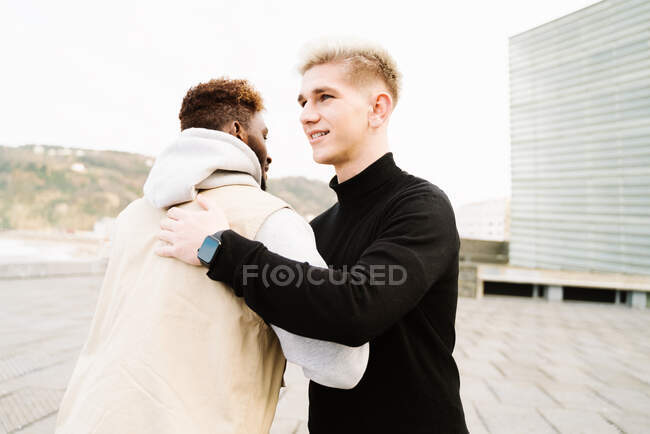 Joyful young multiracial friends in trendy outfits smiling and hugging while standing together on terrace on sunny day — Stock Photo