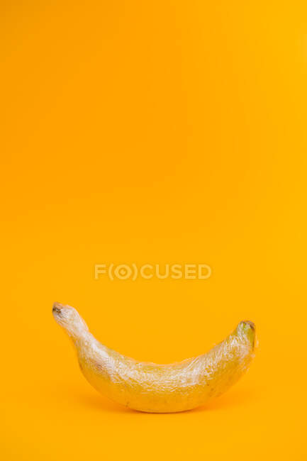 Смачний стиглий банан, покритий прозорою пластиковою обгорткою, що представляє концепцію промислового сільського господарства на яскраво-жовтому тлі — стокове фото