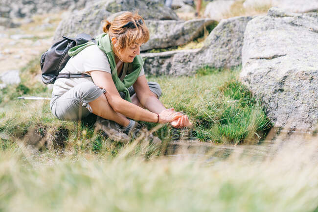 Full body content adult female backpacker in casual wear washing hands in brook while sitting on grassy meadow in stony terrain — Stock Photo