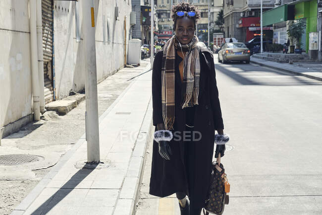 Joven mujer afroamericana de moda con mochila y gafas de sol paseando por la carretera urbana mientras mira la cámara a la luz del sol - foto de stock