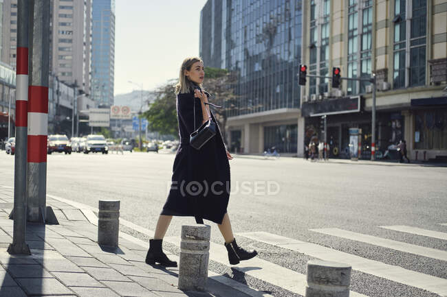 Vista laterale della giovane bionda femmina in abiti neri alla moda passeggiando sul marciapiede in città — Foto stock