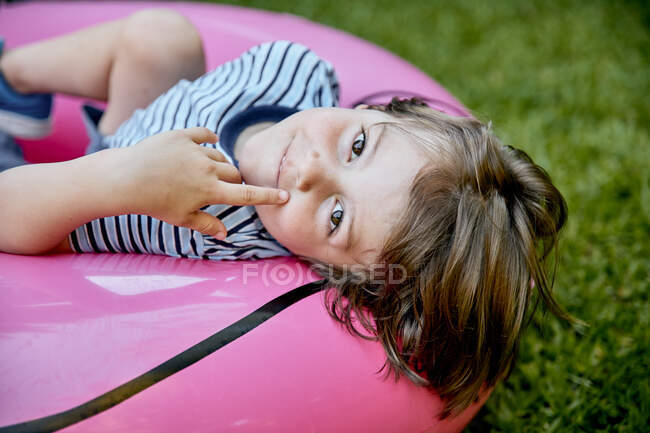 Веселый маленький мальчик в повседневной одежде лежит на надувном розовом фламинго, веселясь на травянистой лужайке в парке — стоковое фото