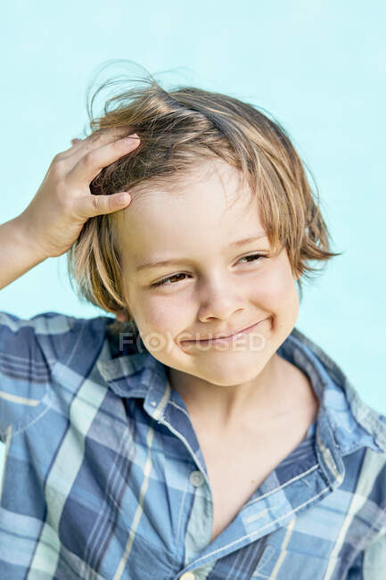 Adorable niño pequeño con el pelo rubio con elegante camisa a cuadros sonriendo y mirando hacia otro lado mientras está de pie sobre el fondo azul a la luz del sol - foto de stock