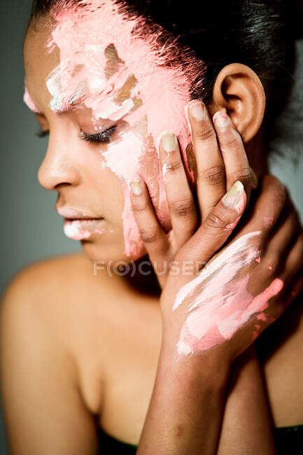 Modèle féminin ethnique créatif avec visage enduit de peinture rose et blanche touchant les joues et regardant loin sur fond gris en studio — Photo de stock