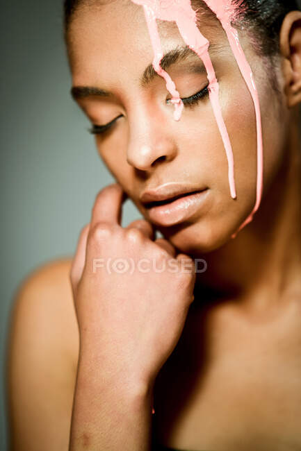Modelo feminino étnico criativo com tinta rosa pingando em seus olhos de rosto fechados em fundo cinza no estúdio — Fotografia de Stock