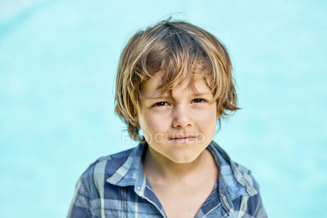 Забавный маленький мальчик с светлыми волосами в клетчатой рубашке, хмурящийся и смотрящий в камеру на синем фоне — стоковое фото
