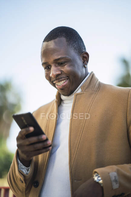 Счастливый молодой афроамериканец в модном наряде на улице и просматривающий мобильный телефон в солнечный летний день — стоковое фото